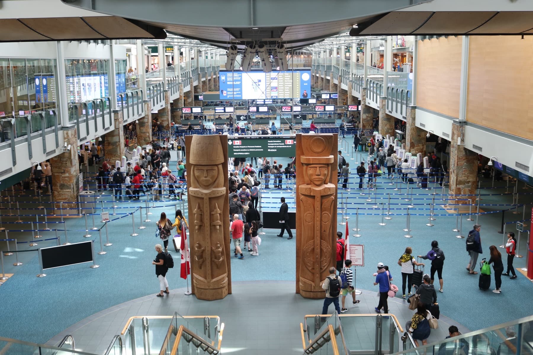 Two carved wooden welcome figures stand in the international arrivals hall at Vancouver International Airport, with travelers queued behind them