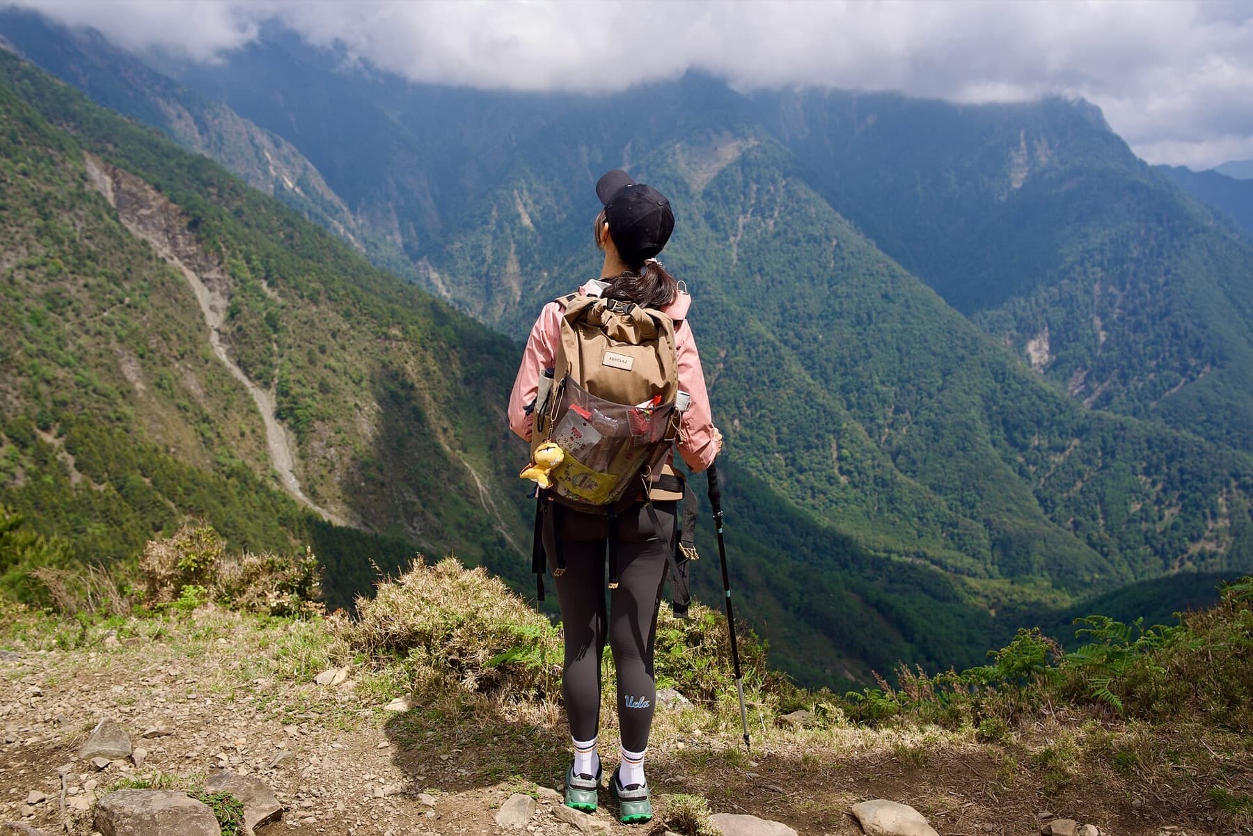 An East Asian hiker with a full backpack and trekking pole stands on a mountain ridge looking across forested peaks