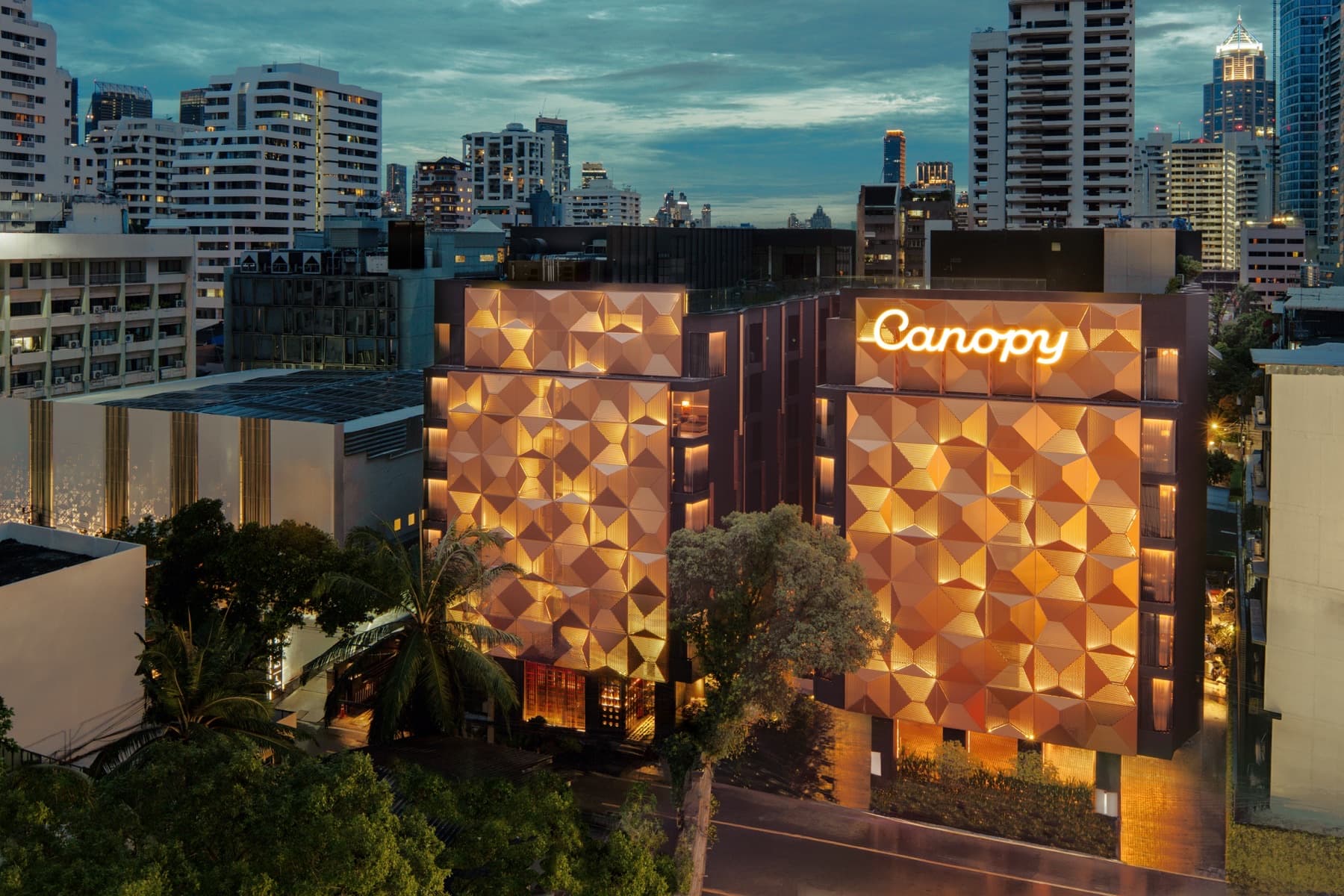 Dusk exterior of Canopy Bangkok Sukhumvit with glowing bronze geometric facade and Canopy signage against the Sukhumvit skyline