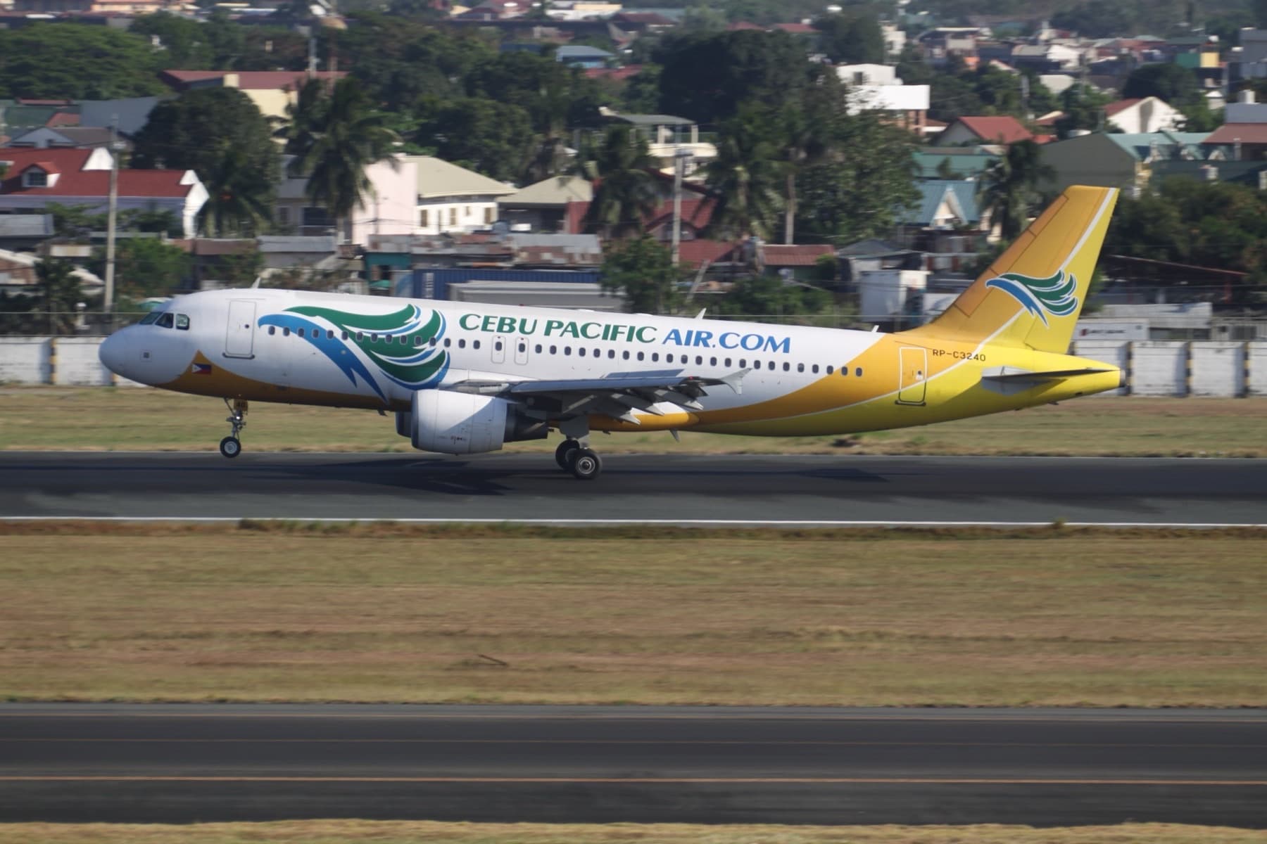 Cebu Pacific Airbus A320 in yellow and green livery on the runway, registration RP-C3240, with palm trees and suburban rooftops beyond the perimeter