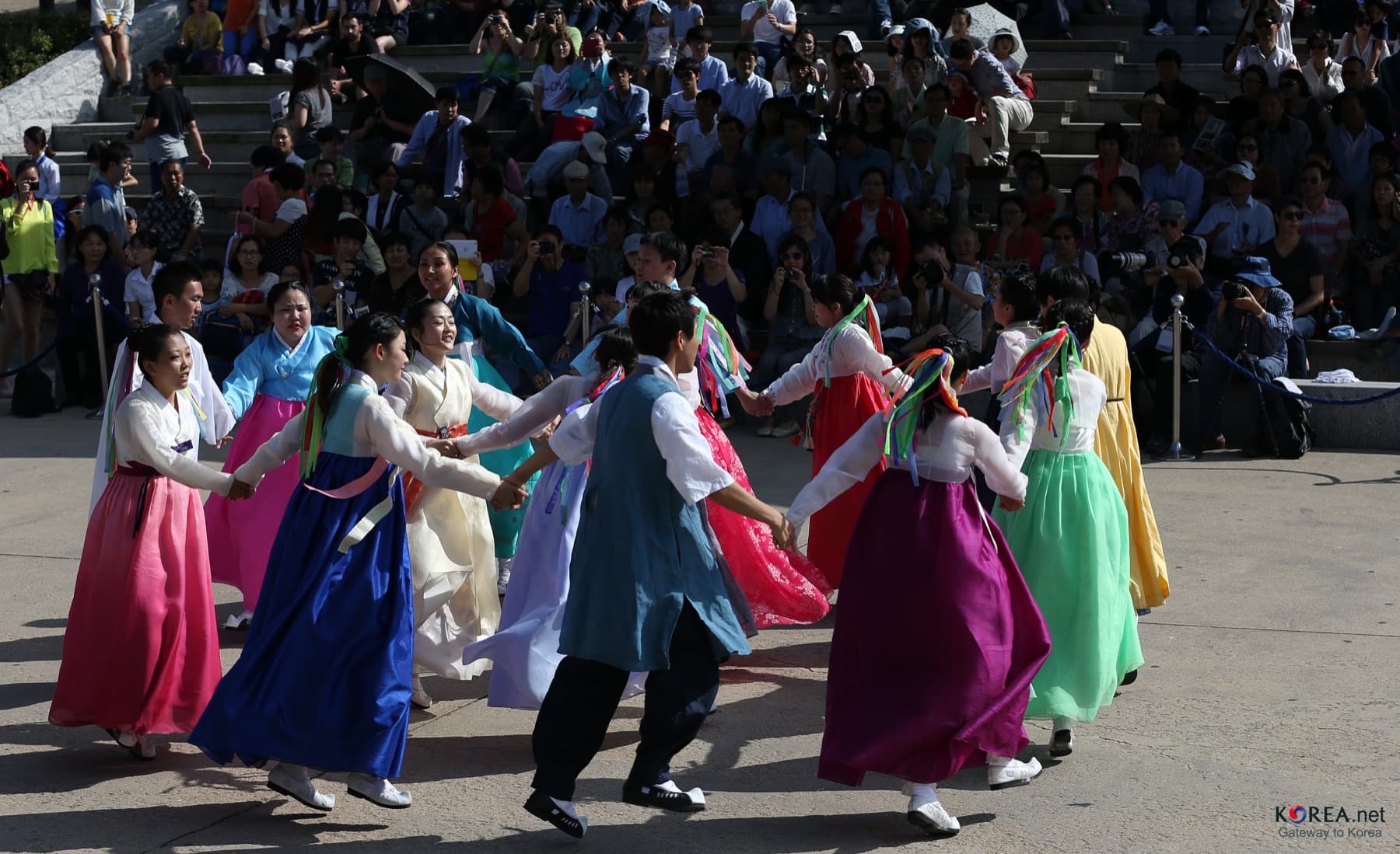 Dancers in colorful hanbok holding hands in a ganggangsullae circle before a crowd on stone bleachers