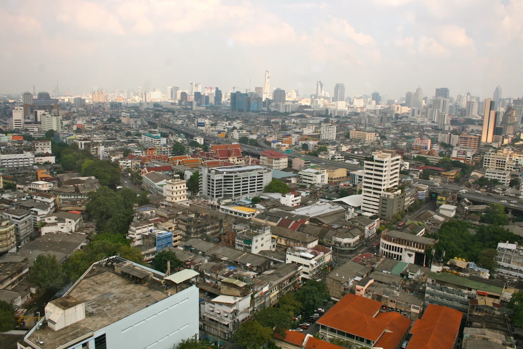 Hazy early-morning Bangkok skyline from a high-floor business hotel, looking over tiled roofs, an elevated highway, and distant towers — The Eight-Day Asia Work Trip Is Long Enough to Exhaust You and Too Short to Adapt