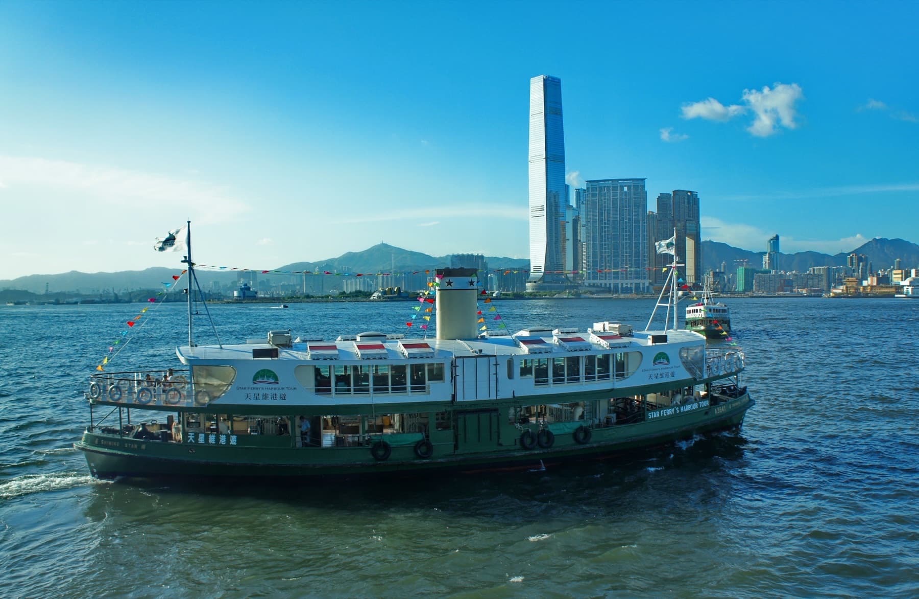 A green-and-white Star Ferry harbour tour boat on Victoria Harbour, with the International Commerce Centre tower rising over West Kowloon behind it
