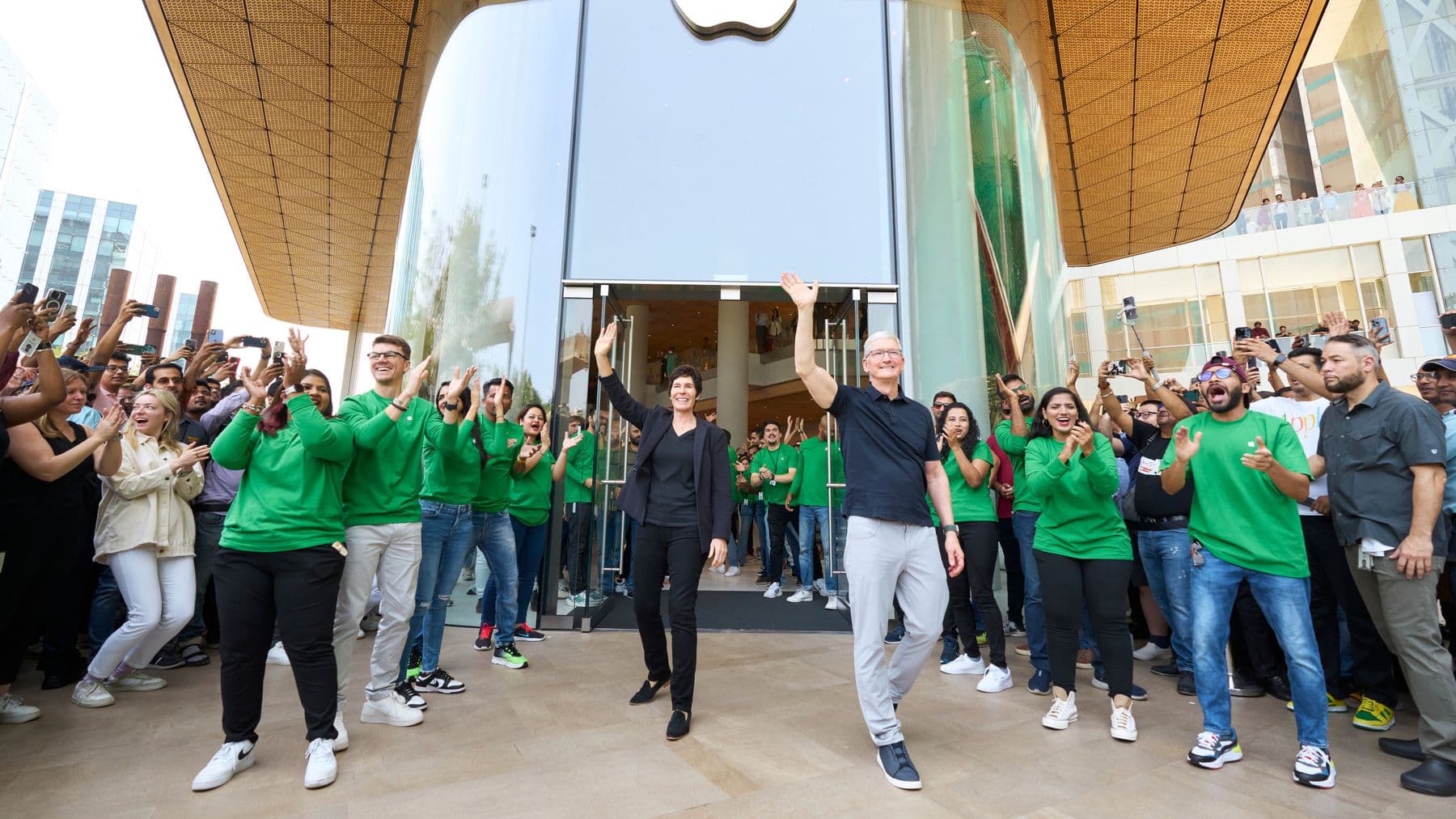 Apple CEO Tim Cook and retail chief Deirdre O’Brien waving outside Apple BKC in Mumbai surrounded by green-shirted team members — India Proposes 2041 Tax Break as Apple Pushes More of Its Supply Chain Beyond China