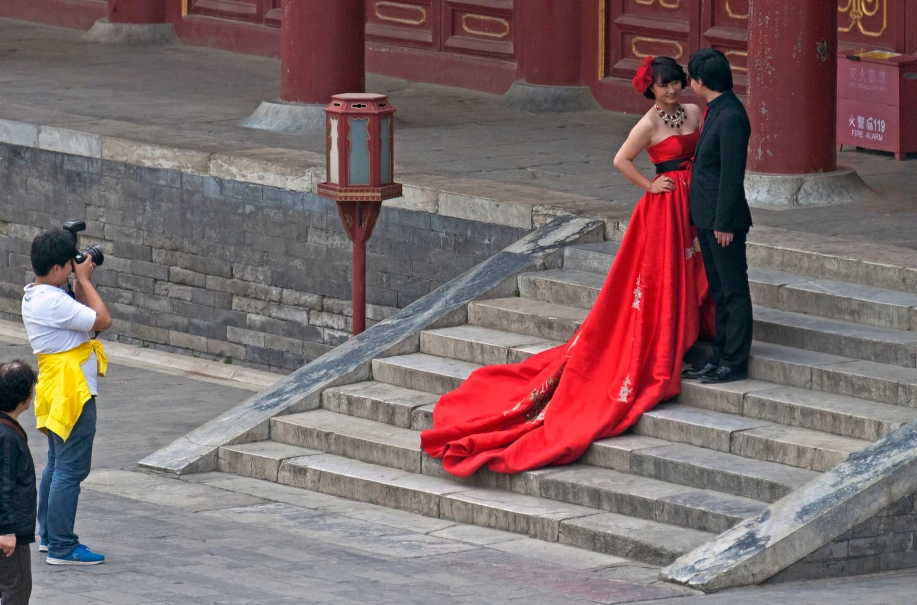 A Chinese couple in a red wedding dress and black suit pose for photographs on the stone steps of a traditional temple building