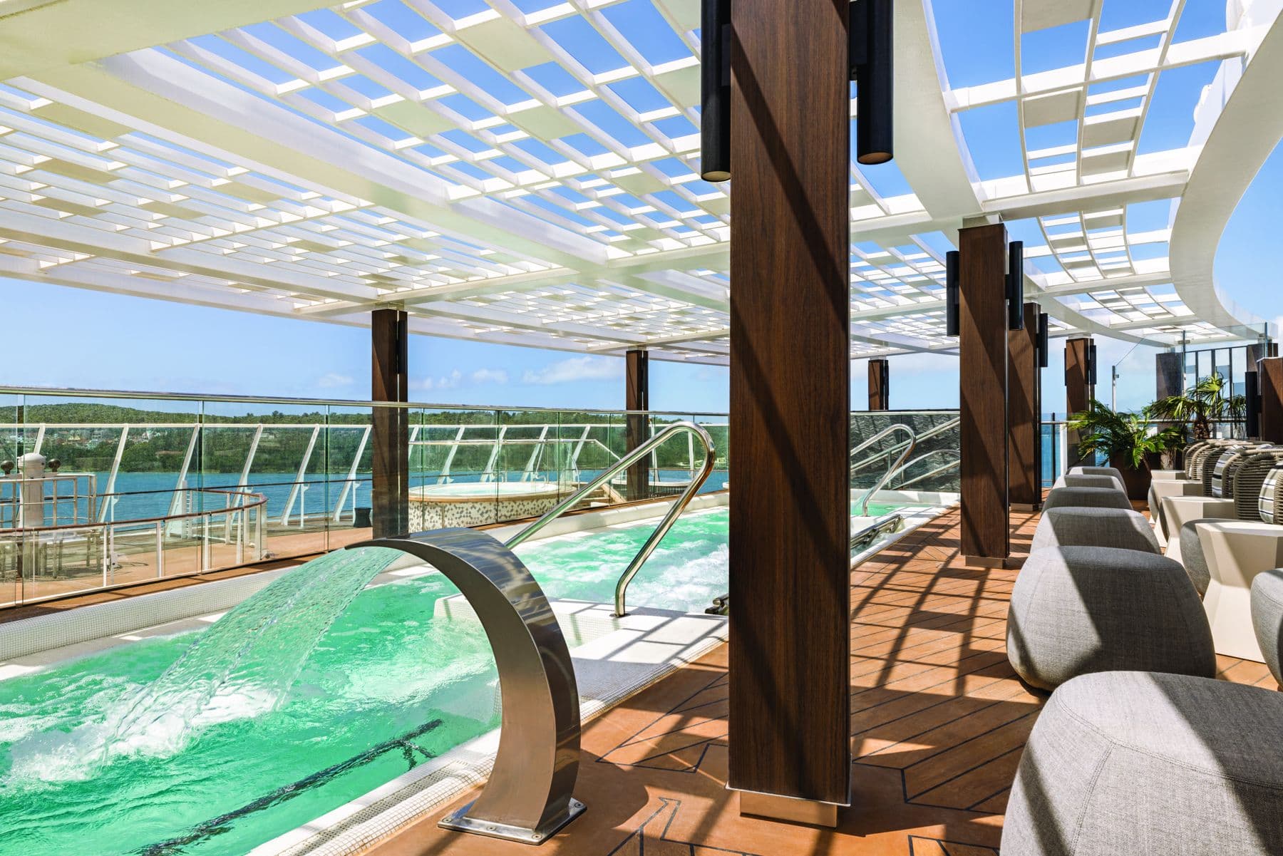 Oceania Cruises Aquamar spa terrace with thermal pool, waterfall spout, and a green coastline beyond the glass railing