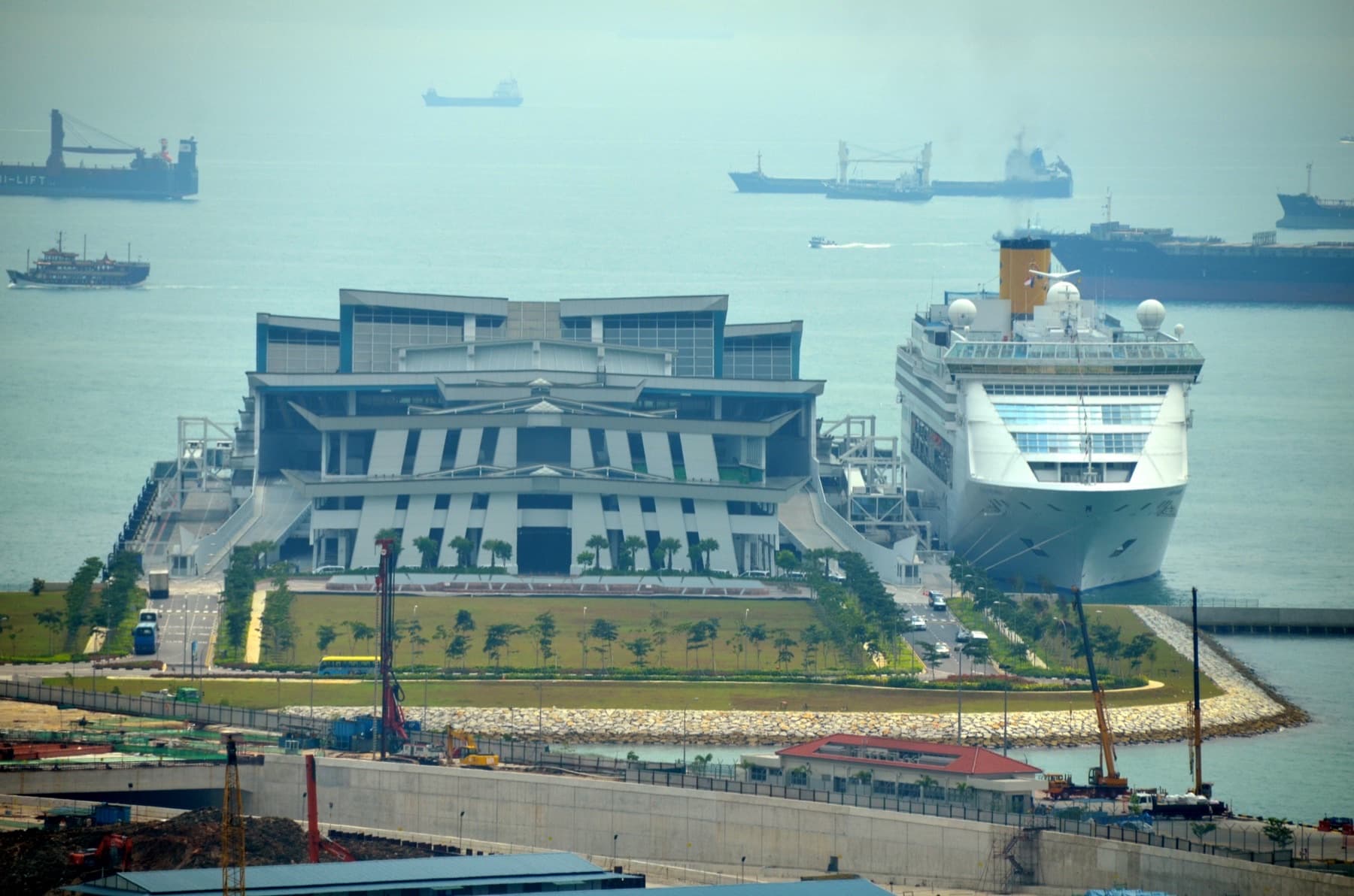 Costa Victoria docked at the angular Marina Bay Cruise Centre Singapore, with shuttle buses on the approach road and cargo ships on the harbor horizon — Beyond Singapore: Southeast Asia Is Building Its Next Generation of Cruise Ports