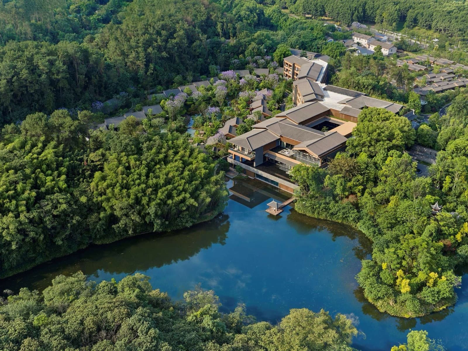 Aerial view of Banyan Tree Mount Emei's tiled-roof buildings beside a lake, set in dense forest at the foot of the mountain