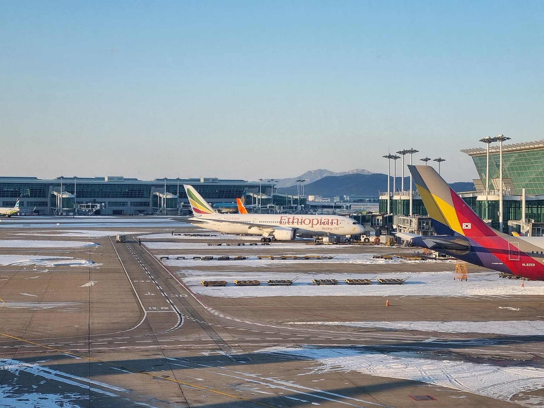 Aircraft on a snowy apron at Incheon International Airport, South Korea, with terminal buildings and mountains beyond